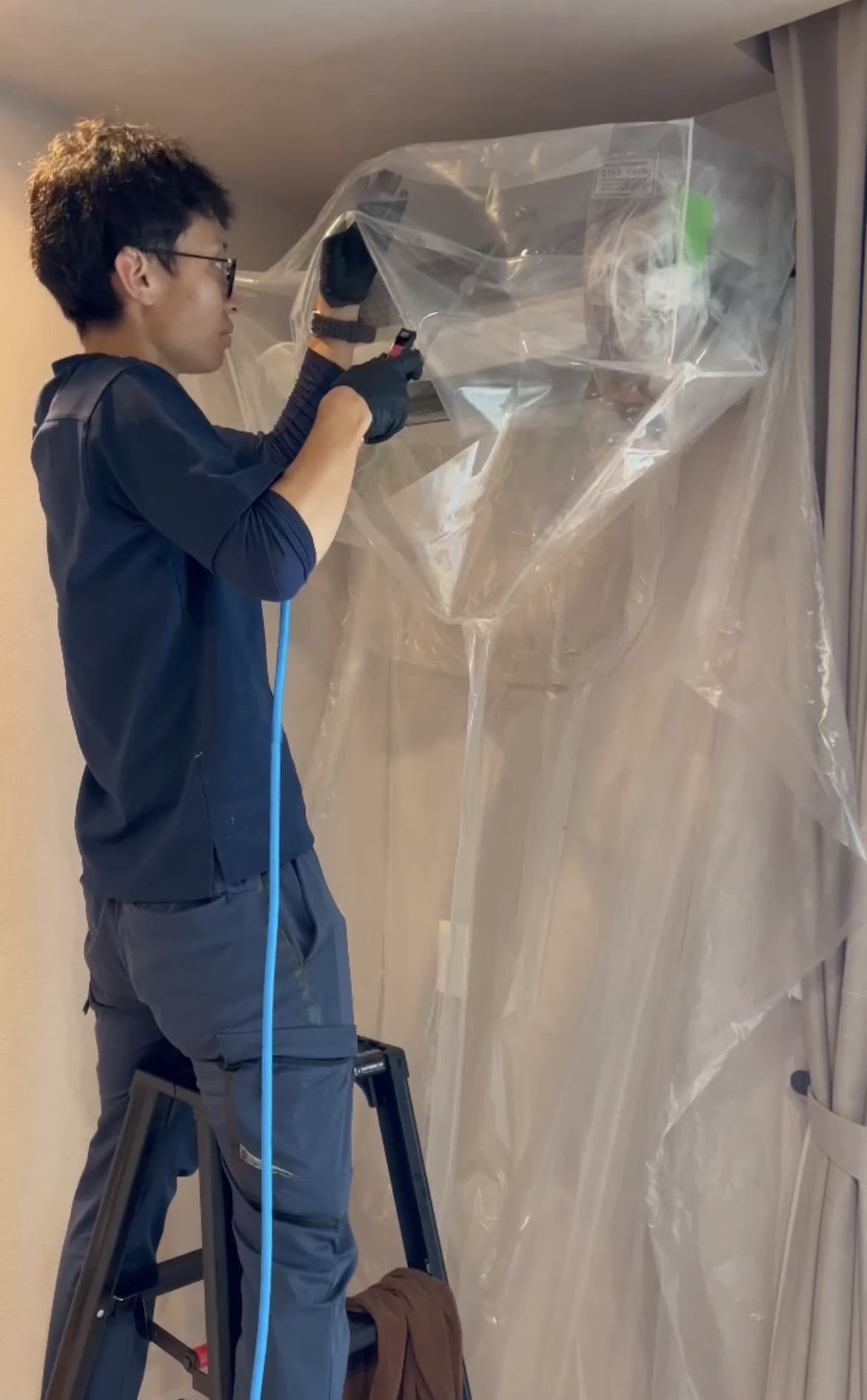 脚立に乗り、エアコン室内機をビニールシートで丁寧に養生するスタッフ（別アングル）